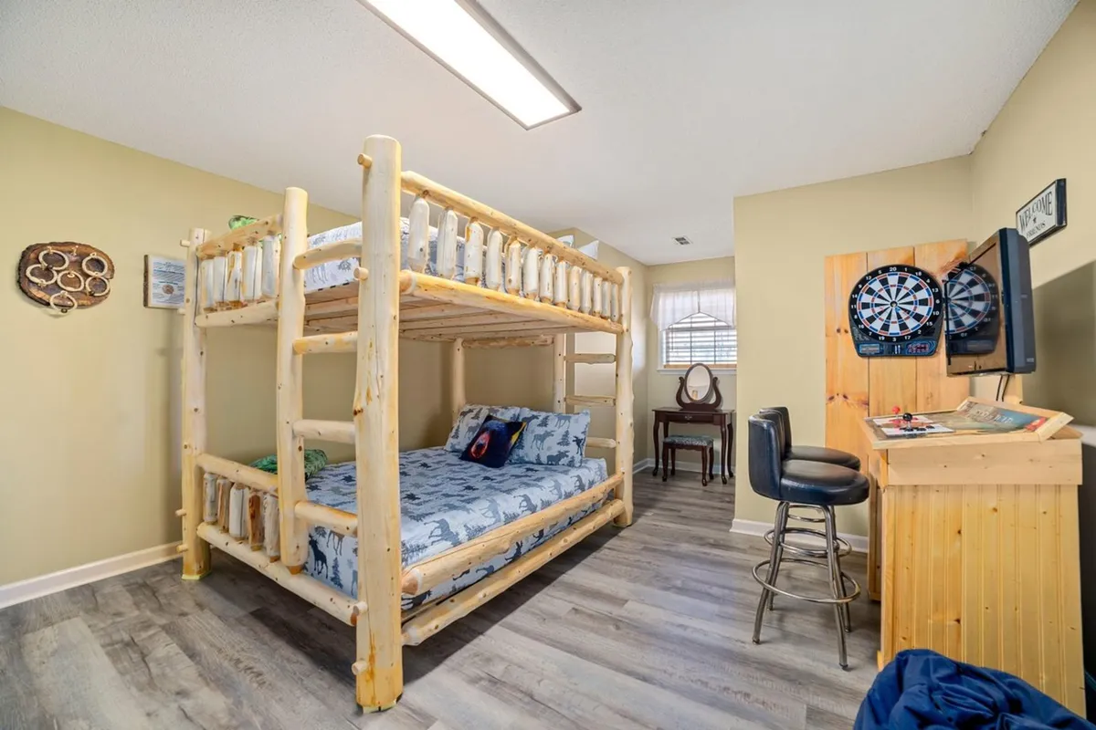 A playful upper-level bunk room equipped with a queen-over-queen bunk bed, a dartboard for games, a wooden desk, and a chair. The space is designed for fun and comfort, making it ideal for children or groups of friends to enjoy both sleep and leisure activities.