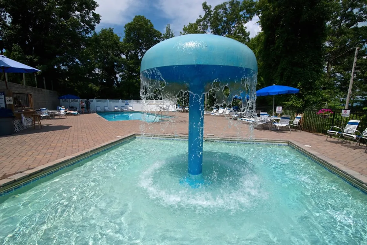 A lively outdoor swimming pool area featuring a water fountain, plenty of lounge chairs, and bright blue umbrellas. The inviting pool is surrounded by greenery and has a playful, energetic atmosphere ideal for families and kids to enjoy a sunny afternoon.