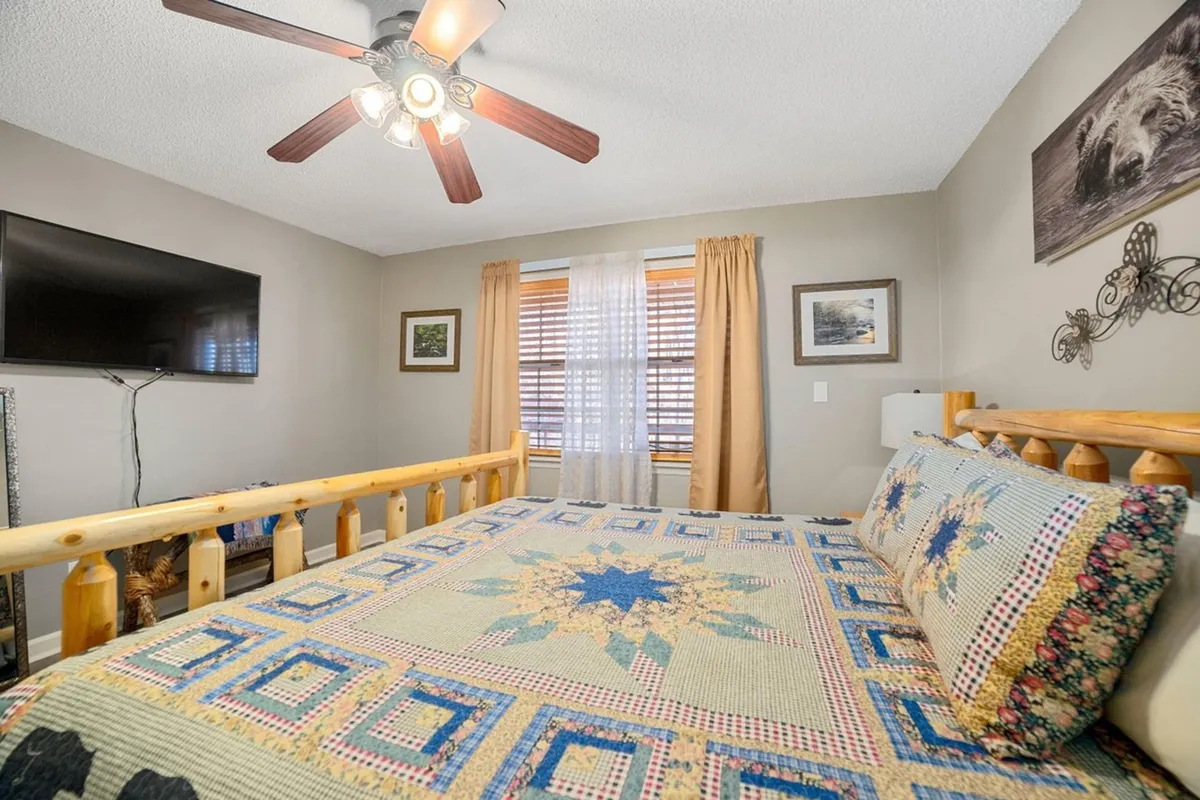 A king bedroom featuring a wooden bed frame with a quilted bedspread, a television mounted on the wall, and a window with soft curtains. The space is bathed in natural light, and the overall feel is warm and homey, perfect for unwinding with entertainment or a good book.
