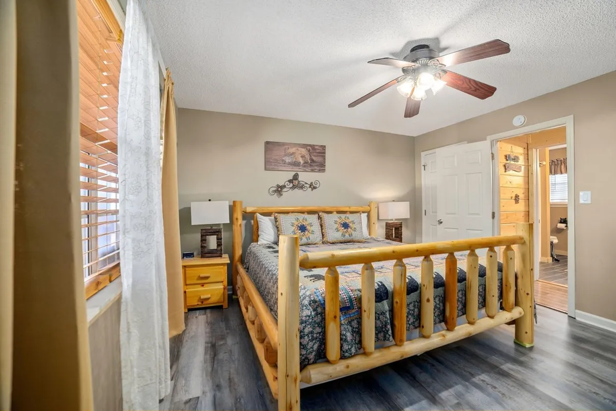 A warmly lit bedroom featuring a rustic log bed adorned with decorative pillows and soft linens. Sunlight streams through the windows, illuminating the cozy space. The room is finished with charming wall art, nightstands, and curtains, creating an inviting environment perfect for rest and relaxation.