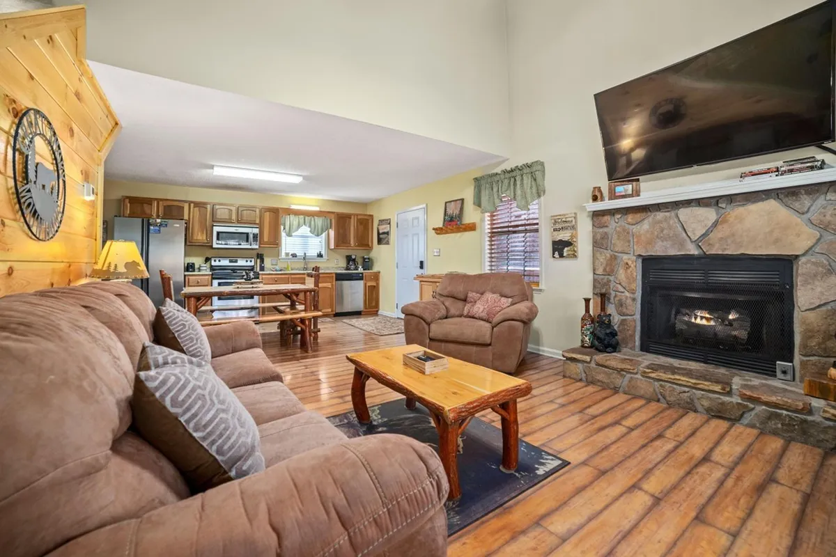 A welcoming, open-concept main living space featuring plush sofas around a glowing fireplace, with a big-screen TV mounted above. The living area seamlessly transitions into a dining space and adjacent kitchen, all within one unified, inviting area—perfect for gatherings, meals, and relaxation.