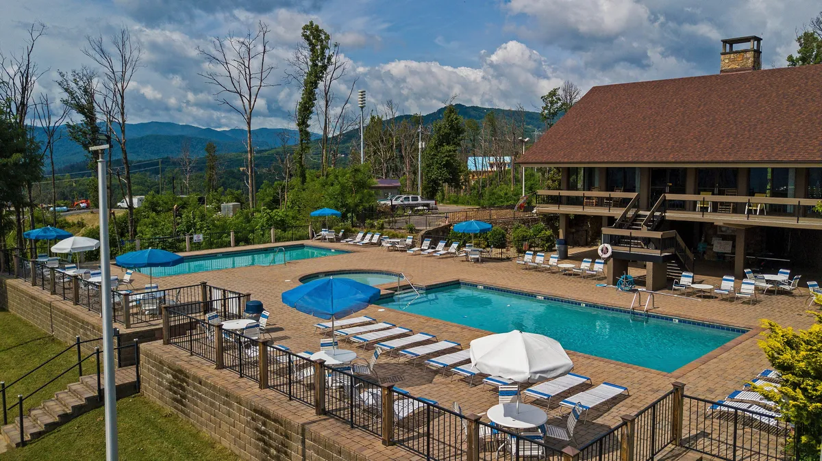 A spacious outdoor pool area with multiple swimming pools, lounge chairs lined with blue umbrellas, and a stunning backdrop of surrounding mountains. The scene captures guests enjoying swimming, sunbathing, and relaxing poolside. The sunset lighting adds a tranquil, vacation-like atmosphere.